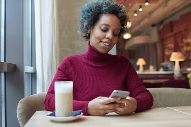 Closeup of beautiful dark skinned woman with colored curly hair spending time alone in cafe holding cellphone in her hands and smiling timidly at content she sees on display with feeling of pleasure. - Photo, Image