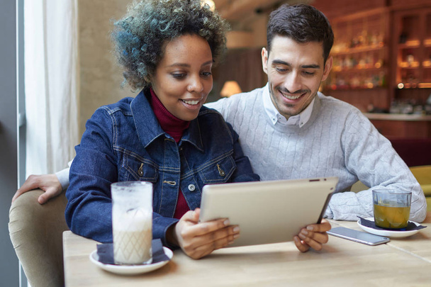 Portrait of joyful smiling interracial couple in cafe, female wearing denim jacket is holding tablet computer and showing pictures and web content to her boyfriend, both looking happily at screen. - Foto, Bild