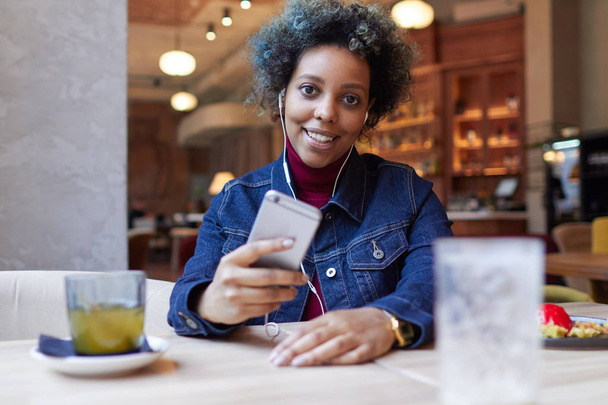 Photo of beautiful smiling African American girl with colored hair spending lunchtime in small cafe on her own dressed casually, holding smartphone and listening to favorite music through earphones. - Fotoğraf, Görsel