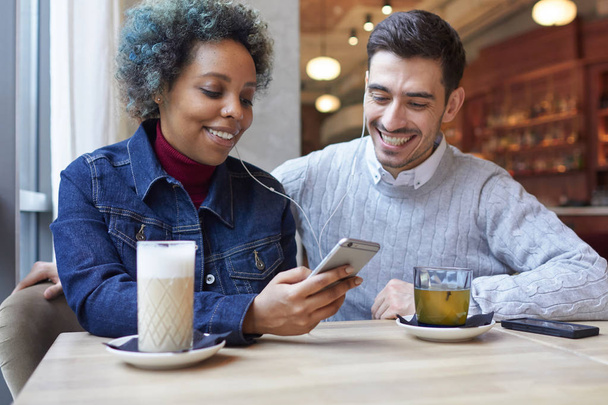 Cheerful dating African American woman and Caucasian man sitting near window in cafe, spending lunchtime together with smartphone earphones on, laughing happily at news from web in atmosphere of joy. - Zdjęcie, obraz