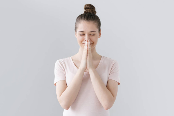 Picture of young girl dressed casually without make up isolated on grey background, having put hands together in prayer or meditation, looking relaxed and calm, dreaming and waiting for all best. - Photo, Image