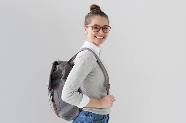 Indoor shot of young European female isolated on gray background standing half-faced with gray bag, wearing trendy eyeglasses, smiling friendly and looking right at camera ready to go where she needs. - Photo, image