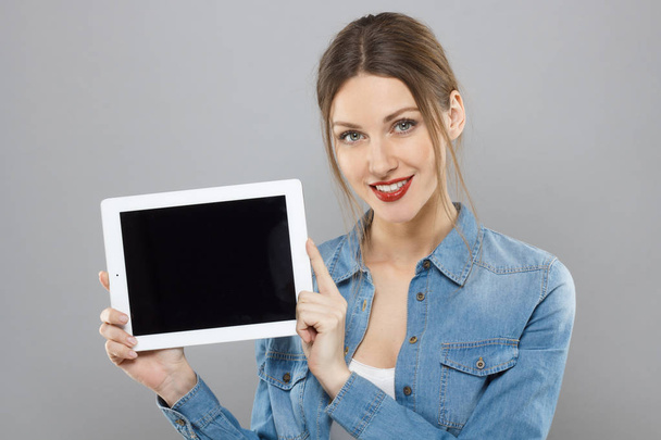 Portrait of young attractive casually dressed girl with red lipstick isolated on gray background smiling gently while showing blank display of tablet computer. Copyspace for advertisement presented. - Fotó, kép