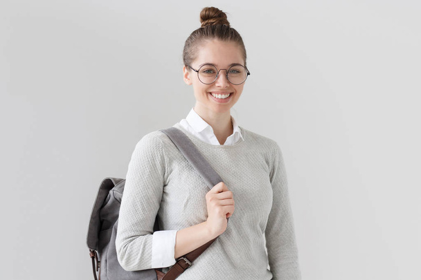 Photo of young European female isolated on grey background, wearing grey trendy backpack and pulling forward it strap, looking straight at camera, smiling and anticipating work or leisure time. - Foto, afbeelding