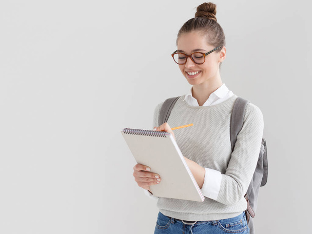 Indoor shot of young European female on right side of photo against gray background taking notes on blank sheets of writing pad she is holding with satisfied and interested smile, happy to study. - Foto, Imagen