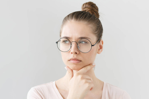 Headshot of good-looking young female in glasses isolated on gray background with head turned leftwards, scratching chin with fingers in deep thinking with uncertain expression and doubt on face. - Foto, immagini