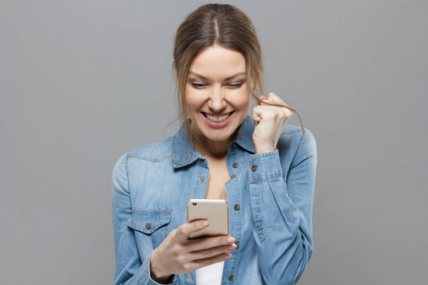 Indoor picture of joyful attractive girl isolated on grey background showing strong positive emotions while holding cellphone in her hand, having received extraordinary great news from web or mates. - Valokuva, kuva
