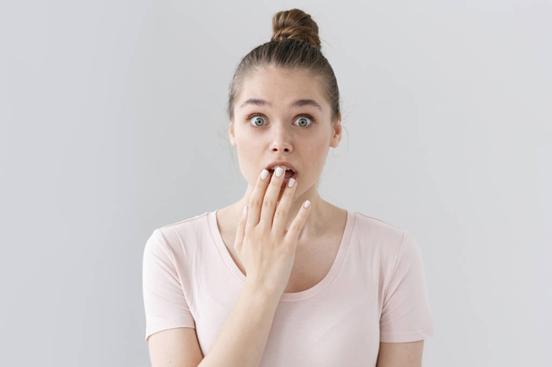 Indoor picture of surprised European female in casual outfit without make up isolated on gray background, looking astonished and deeply shocked, slightly covering round open mouth with hand. - Φωτογραφία, εικόνα