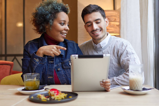 Photo of positive young interracial couple in small restaurant sitting at table together and enjoying using tablet. Girl is pointing to screen, drawing her boyfriends attention to picture.  - Фото, зображення