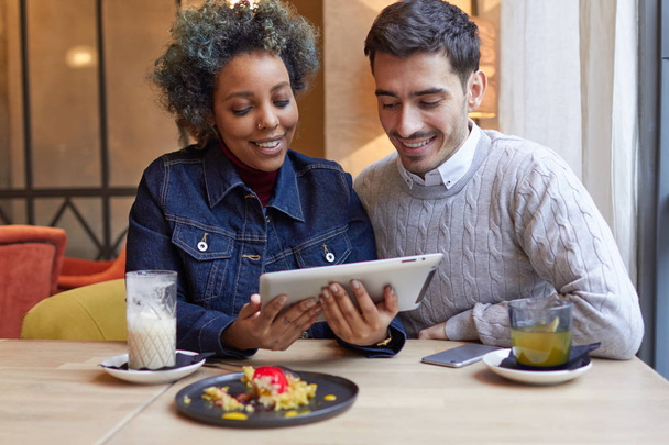 Indoor closeup of young smiling good-looking African American girl and Caucasian guy dressed casually using tablet computer in cafe together browsing Internet and enjoying wireless connection. - Фото, зображення
