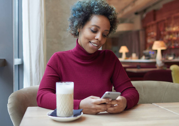 Innenporträt einer gut aussehenden, lässig gekleideten afrikanisch-amerikanischen Dame, die während der Mittagszeit allein im Kaffeehaus sitzt und freundliche Online-Kommunikation per Smartphone mit glücklichem Blick genießt. - Foto, Bild