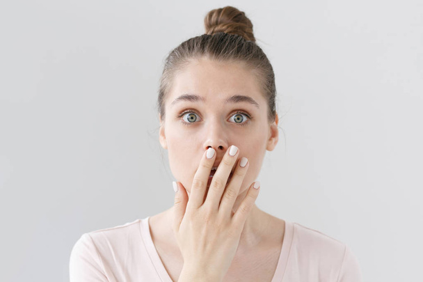 Indoor shot of beautiful teenage girl isolated on gray background dressed in light pink top, covering mouth with hand in sign of deep amazement and shock, hardly believing news she is receiving. - Foto, afbeelding