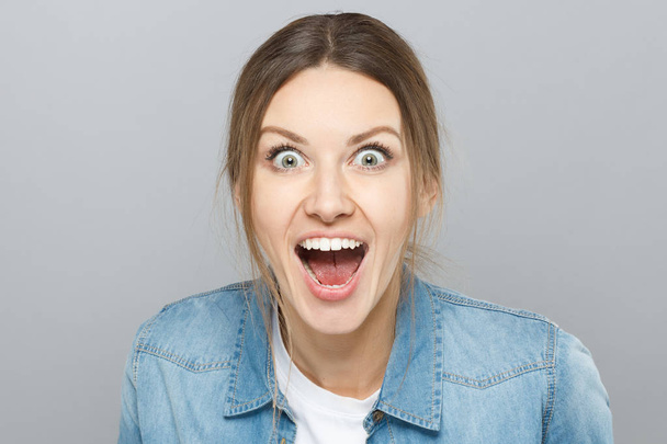 Indoor closeup of extremely emotional European woman isolated on grey background looking crazy with opened eyes, shouting desperately in happiness and euphoria having learnt favorable things. - Foto, Imagem