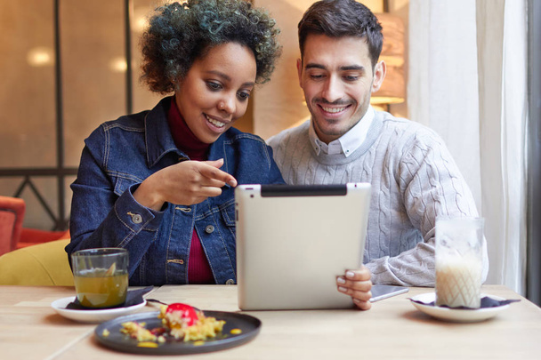 Photo of positive young interracial couple in small restaurant sitting at table together and enjoying using tablet. Girl is pointing to screen, drawing her boyfriends attention to picture.  - Φωτογραφία, εικόνα