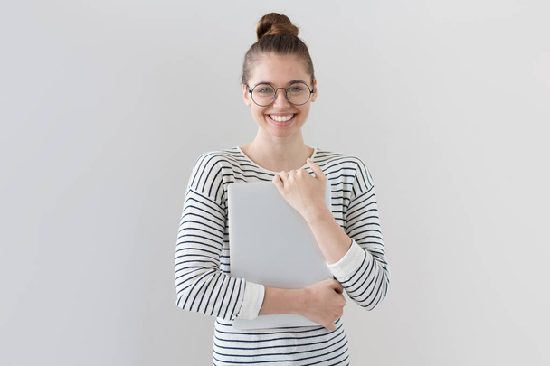 Closeup of young European female isolated on grey background in casual striped top, holding laptop in hands and pressing it to chest with happy smile and expression of enjoyment and satisfaction. - Foto, Bild