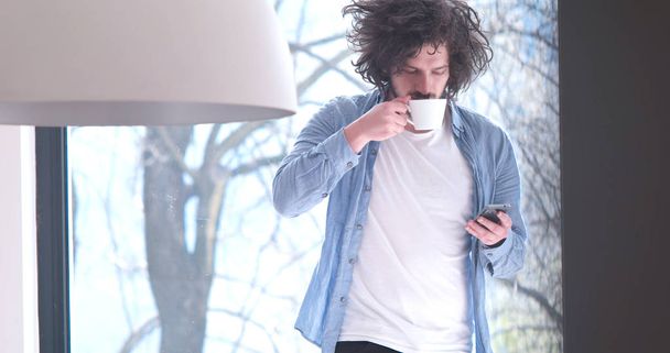 young man drinking coffee and using a mobile phone  at home - Фото, изображение