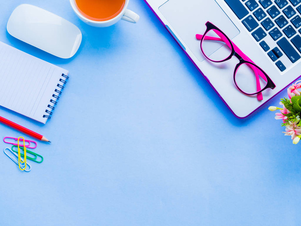 Pohled shora ženský stůl pracovní prostor s kancelářské doplňky včetně notebooku, Poznámka kniha, červenou tužkou, káva, květiny, myší a růžové brýle na modrém pozadí. - Fotografie, Obrázek