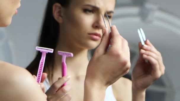 Girl holds a pair of tweezers and a razor, and chooses what to do with epilation - Materiaali, video