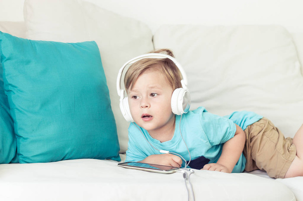 Niño de 3 años se sienta en el sofá y escucha música. Chico lindo viendo dibujos animados en el teléfono. Pequeño niño
. - Foto, Imagen