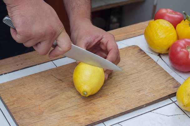 Männliche Hand schneidet mit einem Messer eine reife gelbe Zitrone auf das hölzerne Schneidebrett - Foto, Bild