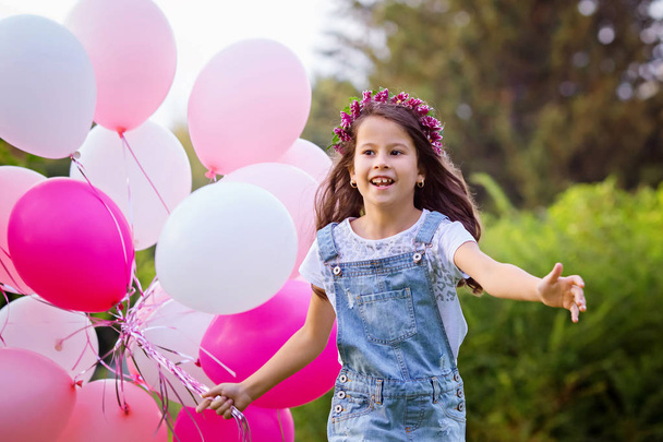Laughing beautiful girl in denim overalls and a floral wreath run to meet with a big bunch of pink balloons. Portrait, selective focus.  - Fotoğraf, Görsel