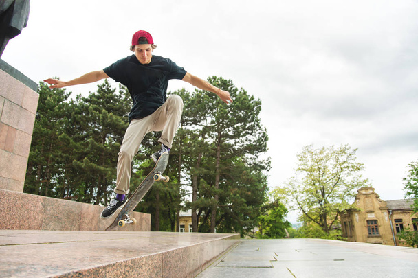 Long-haired skater-teenager in a T-shirt and a sneaker hat jumps an alley against a stormy sky - Foto, afbeelding