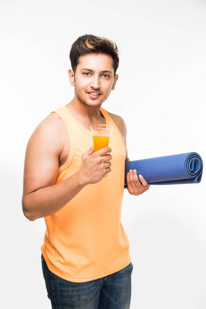 Ψάχνετε καλό Ινδικό νέοι τον άνθρωπο εκμετάλλευση γιόγκα ή άσκηση mat και έχοντας μια γουλιά χυμό σε ένα ποτήρι άσκηση θέση ή γιόγκα / asana, απομονώνονται σε λευκό φόντο - Φωτογραφία, εικόνα