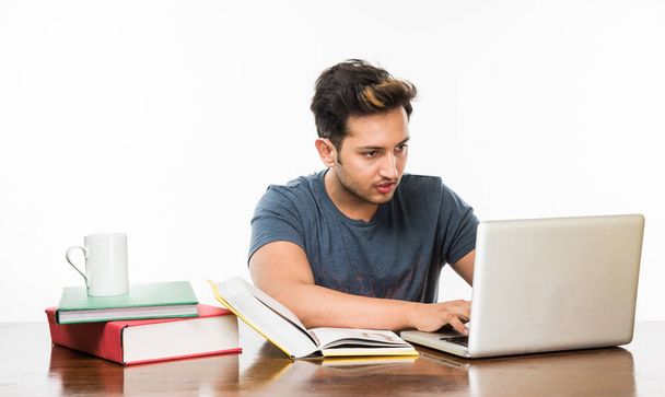 Handsome Indian boy or male college student studying on study table with pile of books, laptop computer and coffee mug. Smiling or thinking or worried or showing thumbs up or using smartphone - Photo, Image