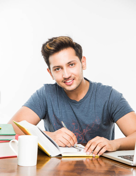 schöner indischer Junge oder männlicher College-Student, der auf einem Arbeitstisch mit Stapel Bücher, Laptop und Kaffeebecher studiert. lächelnd oder denkend oder besorgt oder Daumen hoch oder Smartphone benutzend - Foto, Bild