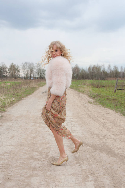 Young blond woman in a beige dress and a short pink fur coat on the countryside road - Foto, Imagen