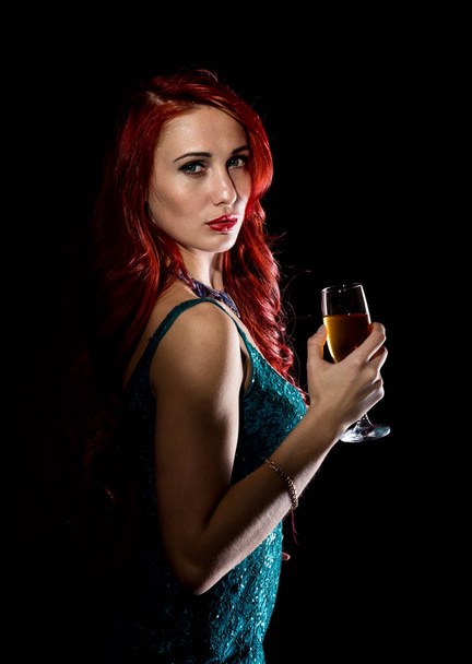 Young redhead sexy woman in blue elegant dress holding wineglass with champagne and poses in a dark. - Foto, imagen