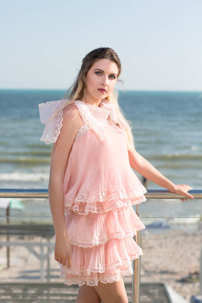 Una bella ragazza posa su un balcone trasparente su uno sfondo blu del mare. Una bella signora in un abito rosa saturo con ruches vicino al mare. Una vacanza estiva godendo
.  - Foto, immagini
