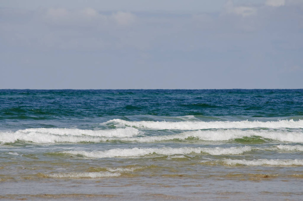 Sandstrand an einem windigen Sommertag mit großen Wasserwellen und hellblauem und bewölktem Himmel - Foto, Bild