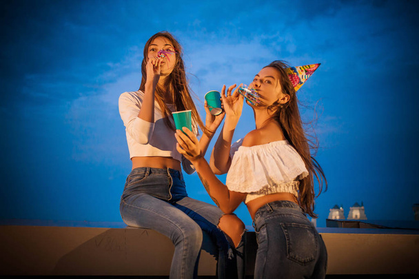 Two attractive young women greet and celebrate the blowing birthday of the trumpet and raise their hands - Fotografie, Obrázek