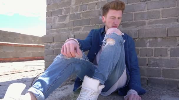 Redhead fashionable young man sitting on the floor of a roof in denim clothes and ripped jeans - Felvétel, videó
