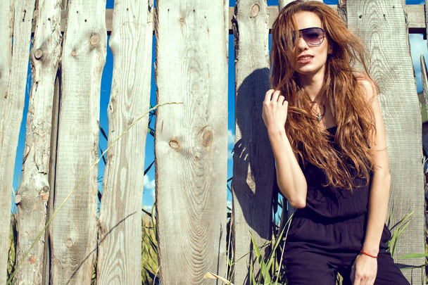 Young beautiful model with long chestnut hair wearing black jumpsuit and trendy aviator sunglasses standing at the old shabby fence made of boards - Zdjęcie, obraz