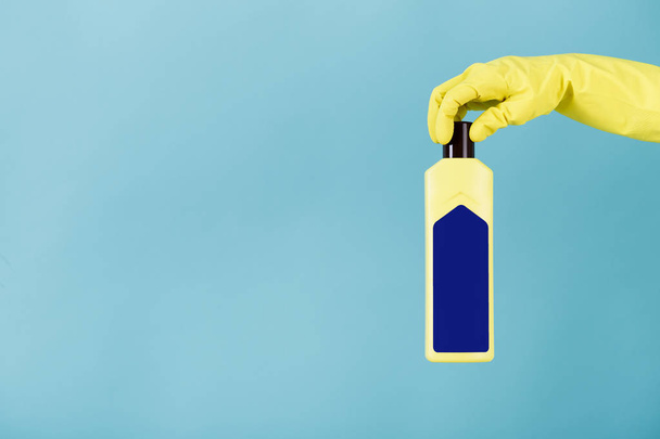 Рука в гумовій жовтій рукавичці тримає пляшку рідкого миючого засобу на синьому фоні. прибирання
 - Фото, зображення