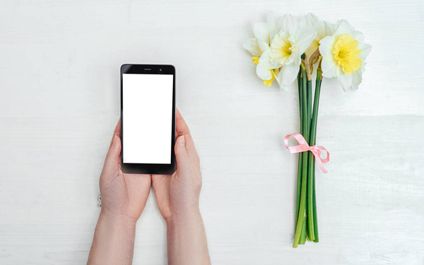 White smartphone screen in the girl's hands near bouquet of narcissus on white background.Technology and communication concept  - 写真・画像