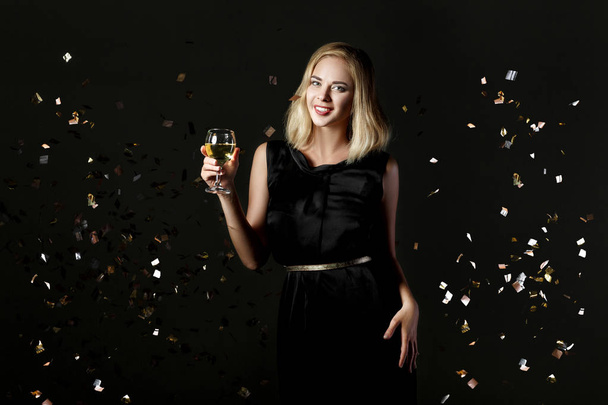 Beautiful happy blonde woman holding glass of white wine on black background with confetti - Foto, imagen