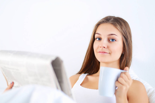 Гарненька молода жінка читає газету в ліжку і насолоджується чашкою чаю
. - Фото, зображення