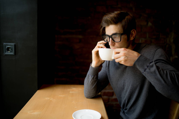 Komea nuori mies puhuu puhelimessa ja hymyilee nykyaikaisessa kahvilassa kahvilla
 - Valokuva, kuva