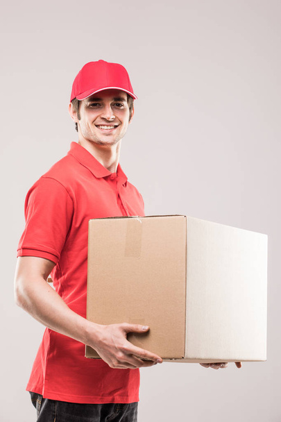 Cheerful delivery man happy young courier holding a cardboard box and smiling while standing on grey - Photo, Image