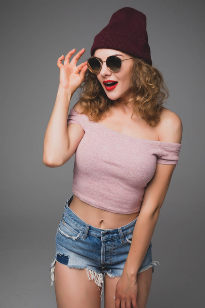 Stylish  fashion portrait of trendy casual young woman in sunglasses and hat on gray background - Foto, imagen