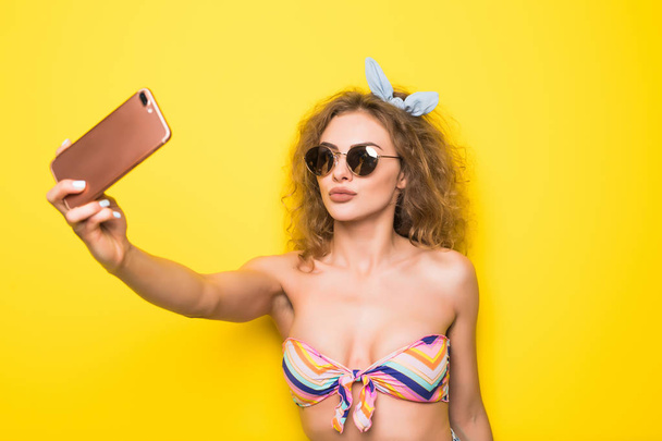 Young beautiful curly woman with happy smile in sunglasses  take selfie on phone  on yellow background - Photo, image