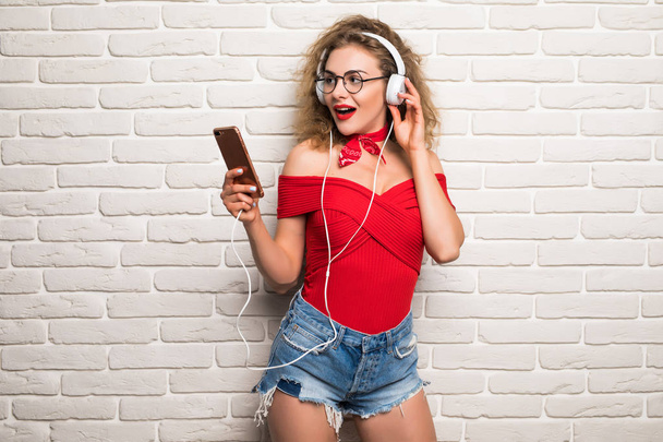 Feliz chica bonita o linda hermosa mujer modelo femenino escucha música en los auriculares contra la pared de ladrillo
 - Foto, Imagen