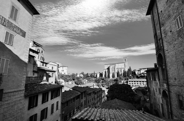 Vista urbana em preto e branco das casas tradicionais em Siena, Itália, com a Basílica de São Domenico ao fundo
 - Foto, Imagem