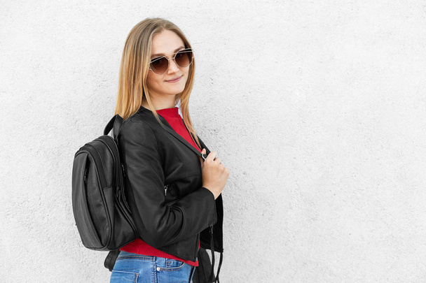 Young female wearing leather black jacket, trendy sunglasses and jeans having rucksack posing against white concrete wall. Female student going to have classes at university posing into camera - Foto, imagen