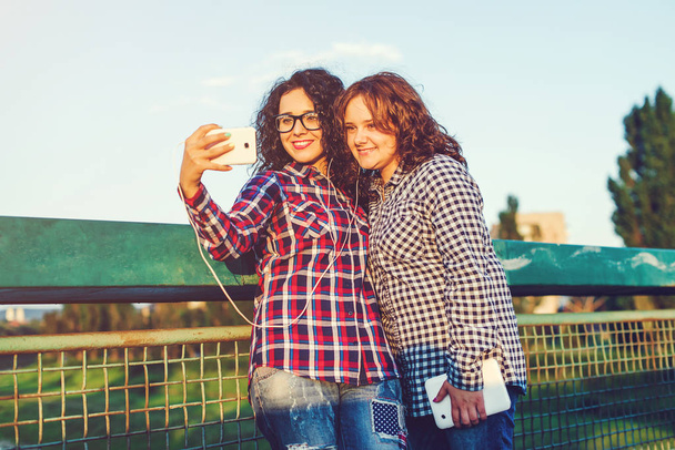 Two girls girlfriend take a selfie on smartphone and listening to music outdoors. - Photo, Image