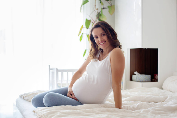 Happy young beautiful pregnant woman, sitting on bed in bedroom - Foto, Imagen