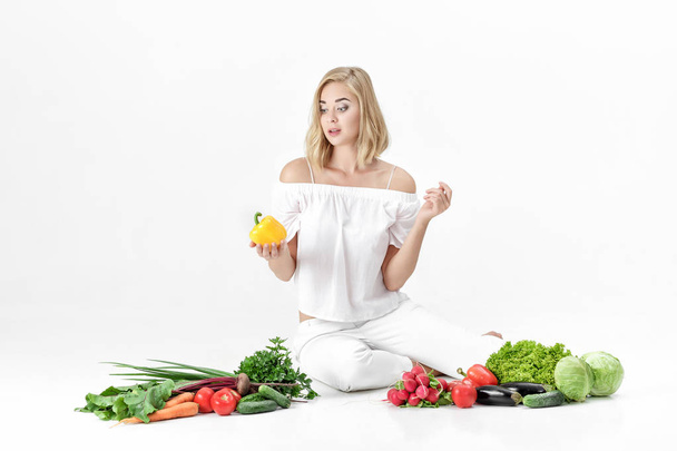 Beautiful blond woman in white clothes and lots of fresh vegetables on white background. Girl holds bell pepper - Fotografie, Obrázek